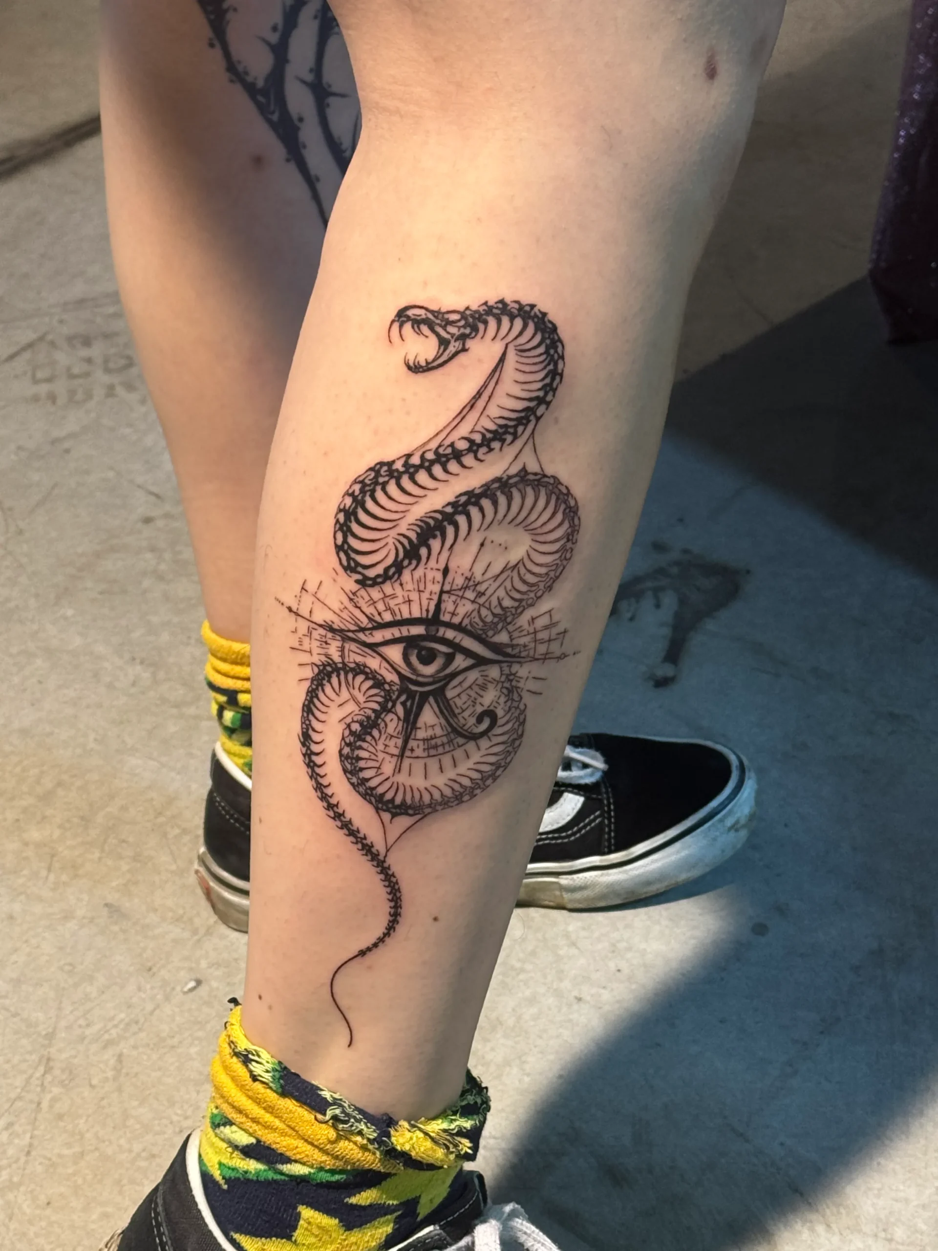 Egyptian-inspired snake tattoo wrapped around the Eye of Horus, created in a detailed black and gray fine line style on the calf.